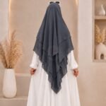 A woman wearing a three layer khimar hijab, with the first layer covering her head and draping down over her chest, and two additional sheer or lightweight layers cascading underneath. BY CLOTHIFYDOTPK