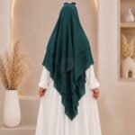A woman wearing a three layer khimar hijab, with the first layer covering her head and draping down over her chest, and two additional sheer or lightweight layers cascading underneath. BY CLOTHIFYDOTPK