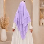 A woman wearing a three layer khimar hijab, with the first layer covering her head and draping down over her chest, and two additional sheer or lightweight layers cascading underneath. BY CLOTHIFYDOTPK