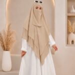 A woman wearing a three layer khimar hijab, with the first layer covering her head and draping down over her chest, and two additional sheer or lightweight layers cascading underneath. BY CLOTHIFYDOTPK