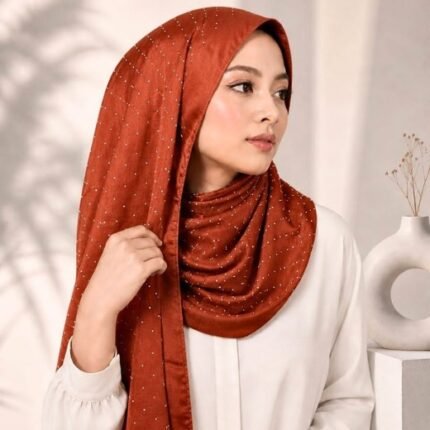 Woman wearing an elegant stone silk hijab in a rust color – soft fabric draping over her head and shoulder with natural lighting.